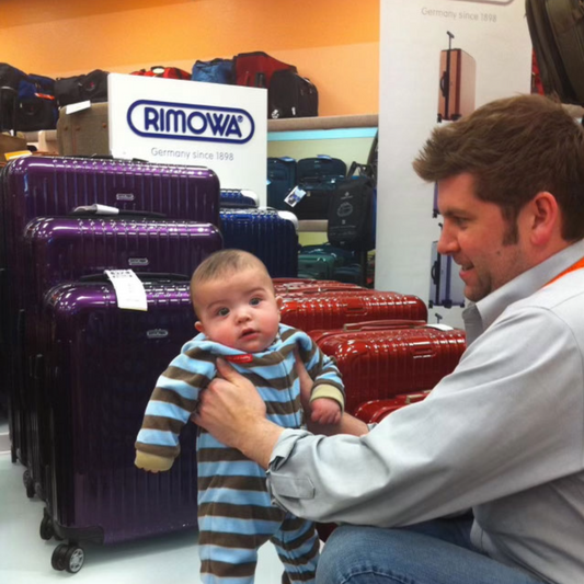Dustin Boyd holding baby Jack in front of Rimowa luggage display at a trade show in 2011